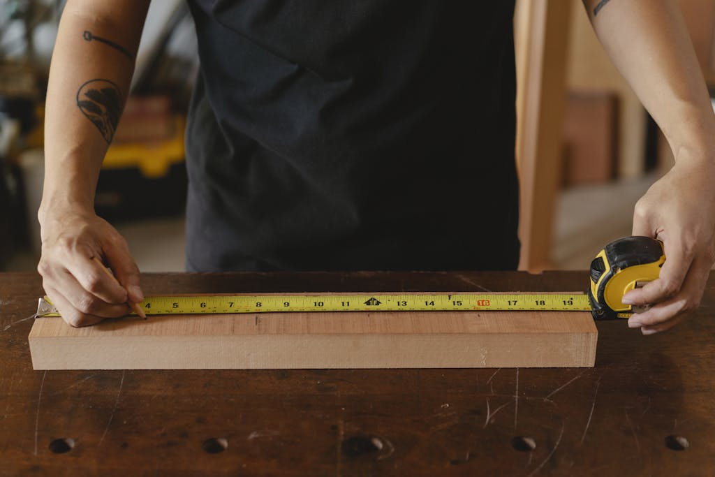 Unrecognizable male carpenter in black outfit with tattoo using ruler on wooden plank while working in studio on blurred background